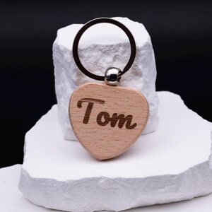 Gravierte Herz Schlüsselanhänger aus Holz mit dem Namen Tom in dekorativer Schrift. Der Anhänger kann individuell personalisiert werden und ist ideal als Geschenkidee für Partner, Freunde oder Kinder. Durch das schlichte, aber moderne Design mit Naturholz und Gravur ist er langlebig und praktisch für den Alltag. Perfekt geeignet für Geburtstage, Jubiläen oder als kleine Aufmerksamkeit.