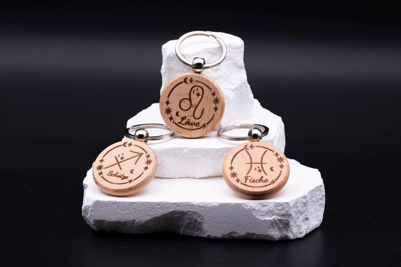 Drei runde Schlüsselanhänger aus Holz mit Sternzeichen-Gravur liegen dekorativ auf weißem Steinpodest vor schwarzem Hintergrund. Sichtbar sind die Sternzeichen Löwe, Schütze und Fische, jeweils mit astrologischem Symbol, Sternenkranz und Schriftzug in geschwungener Schrift. Die Anhänger haben einen silberfarbenen Metallring zur Befestigung am Schlüsselbund. Das natürliche Holz und die feine Gravur betonen die handwerkliche Qualität und den personalisierten Charakter.