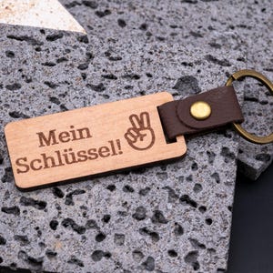 Hochwertiger Schlüsselanhänger aus Holz mit Gravur „Mein Schlüssel!“ und Symbol der Victory-Hand. Der rechteckige Holzanhänger ist mit einem braunen Kunstlederband und stabilem Messingring versehen. Praktisch, langlebig und individuell gravierbar – ideal als personalisiertes Geschenk für Männer, Frauen oder Freunde. Modernes Design kombiniert natürliche Holzoptik mit edlem Lederlook für Alltag und besondere Anlässe.