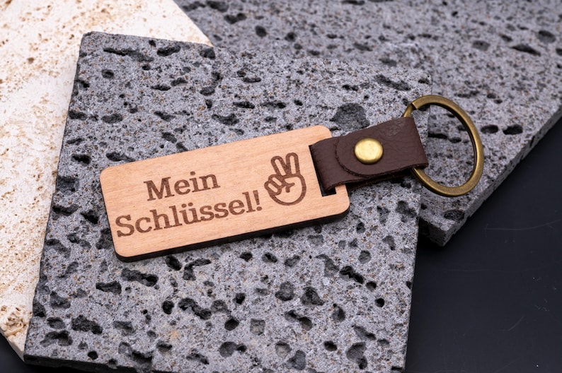 Hochwertiger Schlüsselanhänger aus Holz mit Gravur „Mein Schlüssel!“ und Symbol der Victory-Hand. Der rechteckige Holzanhänger ist mit einem braunen Kunstlederband und stabilem Messingring versehen. Praktisch, langlebig und individuell gravierbar – ideal als personalisiertes Geschenk für Männer, Frauen oder Freunde. Modernes Design kombiniert natürliche Holzoptik mit edlem Lederlook für Alltag und besondere Anlässe.