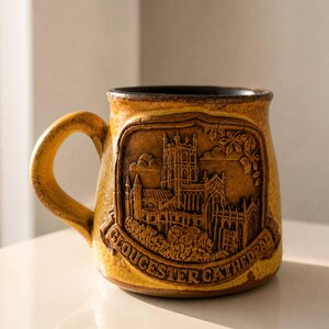 May include: A ceramic mug with a detailed relief of Gloucester Cathedral. The mug is a warm, earthy yellow-brown color with a black interior. The handle is a matching yellow-brown. The words "Gloucester Cathedral" are embossed on the mug.