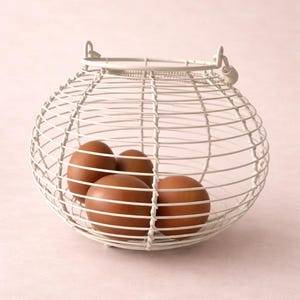 May include: A white wire egg basket containing four brown eggs. The basket is spherical with a handle and sits against a soft pink backdrop. The wire design allows for easy viewing of the eggs.