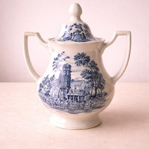 Puede incluir: Azucarero de porcelana blanca con tapa y dos asas. El cuenco y la tapa están decorados con una escena azul y blanca de transferencia de un edificio, árboles y otros elementos arquitectónicos. El azucarero tiene una base de pedestal.