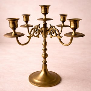 Puede incluir: Un candelabro de latón con cinco candeleros. El candelabro tiene una base redonda, un tallo central y brazos curvos que se extienden para sostener las velas. El acabado es de color oro antiguo cálido.