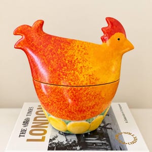 Puede incluir: Un recipiente de cerámica con forma de gallina que presenta un degradado vibrante de amarillo, naranja y rojo con un acabado texturizado. La base está decorada con un patrón de círculos amarillos. El recipiente descansa sobre un libro titulado The Times Picture Collection London.
