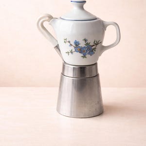 May include: A white ceramic coffee pot with a blue floral pattern sits on a silver metal coffee maker. The ceramic pot has a lid and handle, and the metal base has a conical shape. The background is a soft, neutral colour.