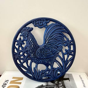 Puede incluir: Un salvamanteles circular de cerámica azul oscuro brillante con un diseño en relieve de un gallo de pie entre flores y follaje estilizados. El gallo está representado de perfil, mirando hacia la izquierda, con plumas detalladas.