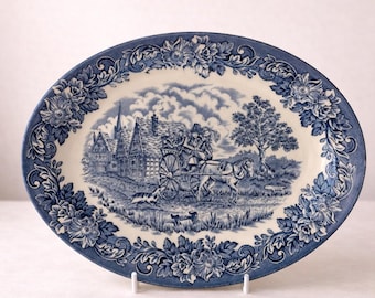 May include: Oval blue and white ceramic platter with a floral border. The center features a pastoral scene with a horse-drawn carriage, houses, and trees. A decorative serving dish for displaying food or as a decorative piece.