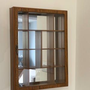 Pode incluir: Um armário de exibição de madeira montado na parede com uma porta de vidro e várias prateleiras. O armário tem uma forma retangular com um acabamento marrom e uma alça decorativa. O interior é dividido em vários compartimentos.