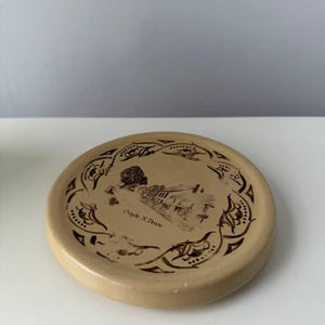 Puede incluir: Un posavasos de cerámica beige con una ilustración marrón de un edificio y un árbol, rodeada por un borde decorativo. El texto "Creede X Devon" está impreso debajo. El posavasos es redondo.