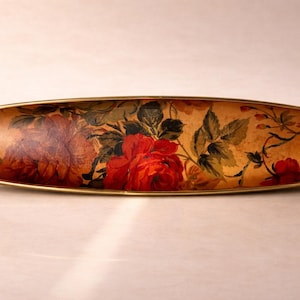May include: An oval decorative tray with a floral design. It has a gold-coloured metal rim and a painted surface featuring red roses, green leaves, and a tan background. The tray is a decorative item, suitable for display.