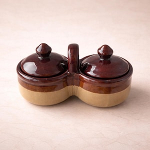 May include: A two-section ceramic condiment server with matching lids and a handle. The server is brown with a tan base. The lids have a rounded knob. The server is designed for serving condiments.