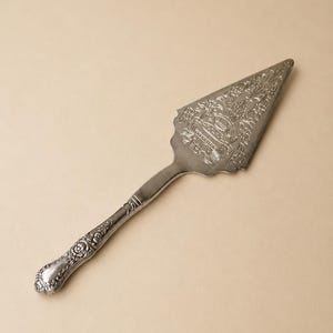 Może przedstawiać: Srebrzysta metalowa łopatka do ciasta z ozdobnym, tłoczonym wzorem kwiatowym i roślinnym na trójkątnym ostrzu oraz pasującym dekoracyjnym uchwytem z misternymi detalami wzdłuż krawędzi i ząbkowaną krawędzią tnącą po prawej stronie.