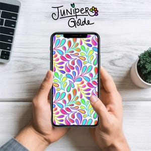 May include: A smartphone displays a colorful, abstract pattern of stylized leaves in shades of red, orange, yellow, green, blue, and purple. The phone is held in two hands against a white wood-grain background. The text "Juniper Golde" is at the top.