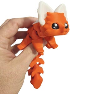 May include: A 3D-printed orange articulated fox toy with white ears and large black eyes. The fox is held in a hand, showcasing its poseable limbs and tail. The toy is designed with a cute, cartoonish aesthetic.