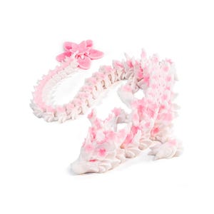 May include: A white articulated dragon figurine featuring pink accents along its spine and head. The dragon has a long, segmented body and a tail that ends in a five-petaled pink flower. The surface has a matte texture throughout the design.