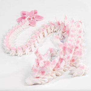 May include: A white and pink dragon sculpture with a flower-shaped accent. The dragon is detailed with a spine and flower-like accents in pink. The sculpture is on a white background.