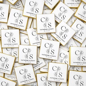 May include: Square white favors with gold edges, featuring the initials "C" and "S" with a floral design, and the text "CATHERINE & SCOTT 05.10.23". These personalized wedding favors are scattered across a surface.