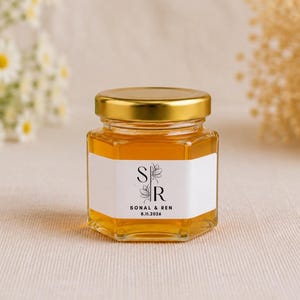 May include: A small hexagonal glass jar filled with golden honey, featuring a metallic gold lid and a white label. The label displays the letters S and R in a stylized font, along with the text SONAL & REN and the date 8.11.2026 in black ink.