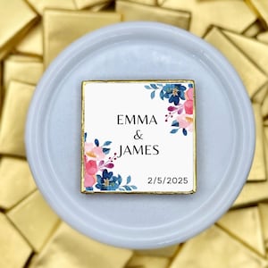 Op de afbeelding: Een vierkant chocoladebedankje met gouden rand, met de namen "EMMA & JAMES" en de datum "2/5/2025", omgeven door roze en blauwe bloemaccenten. Het bedankje ligt op een lichtblauw bord, met goud verpakte chocolaatjes op de achtergrond.