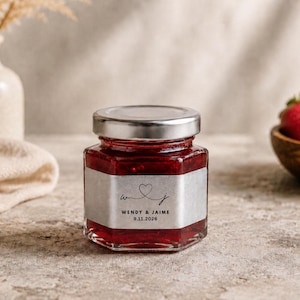 May include: A hexagonal glass jar filled with bright red strawberry jam, featuring a silver label with a heart icon, the initials W and J, and the text WENDY & JAIME 9.11.2026. The jar has a metallic silver screw-top lid and clear glass sides.