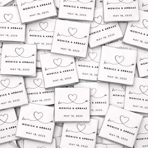 May include: A collection of white square favors, each with a heart design and the names "MONICA & ARBAAZ" along with the date "MAY 18, 2025" printed in black. The favors are scattered, creating a visually appealing arrangement.