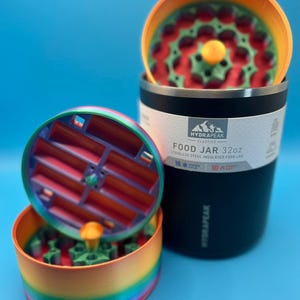 May include: A black 32oz stainless steel insulated food jar with a rainbow-colored lid and a matching rainbow-colored food container. The lid is open, revealing a red and green interior. The jar has the text "HYDRAPEAK" and "FOOD JAR 32oz".