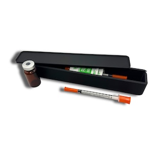 May include: A black rectangular case with a lid, containing medical supplies. An amber vial with a silver cap sits outside the case. A syringe with an orange cap and a roll of alcohol prep pads are also visible.