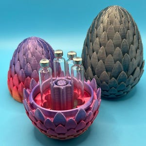 May include: A 3D-printed, multi-colored egg-shaped container with a flower-like design, holding several small glass vials. A larger, gray egg-shaped object is behind it. The objects are on a blue surface.