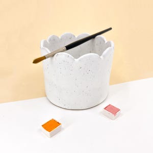 Pode incluir: Prato de pintura de cerâmica branca com uma borda festonada e um esmalte salpicado. Um pincel repousa na borda do prato. Duas pastilhas de tinta aquarela estão sobre uma superfície branca.