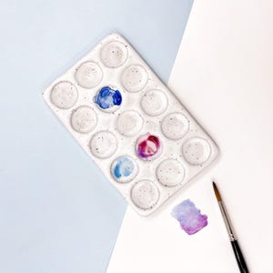 Könnte beinhalten: Weiße Keramik-Aquarellpalette mit 18 runden Vertiefungen. Die Palette hat eine gesprenkelte Textur und ist mit blauer, rosa und violetter Aquarellfarbe gefüllt. Ein Pinsel liegt neben der Palette auf einer weißen Oberfläche.