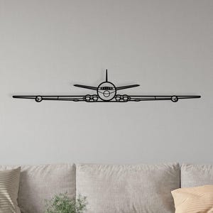 Op de afbeelding: Zwarte metalen vliegtuig wanddecoratie. Het kunstwerk toont een vooraanzicht van een vliegtuig met uitgespreide vleugels. Het is gemonteerd op een lichtgrijze muur boven een bank en salontafel, wat een decoratief element met luchtvaartthema biedt.