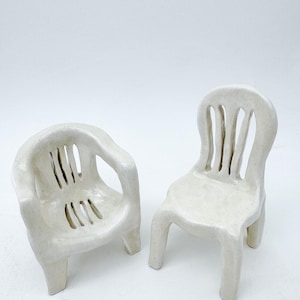Peut inclure: Deux chaises en céramique blanche au design simple et moderne. Les chaises ont des dossiers ouverts et sont réalisées dans un style légèrement abstrait.