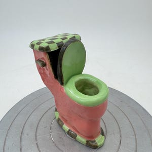 Puede incluir: Una pequeña figurita de inodoro de cerámica con una tapa a cuadros verde y negro, un asiento verde y una base rosa.