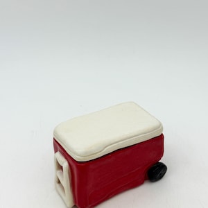 Puede incluir: Una pequeña nevera roja con tapa blanca y una sola rueda negra. La nevera tiene un asa lateral. El diseño es una réplica en miniatura de una nevera real. El tema es la decoración en miniatura.