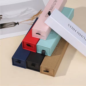 Puede incluir: Una colección de cajas rectangulares coloridas con cajones extraíbles. Las cajas son de varios colores, incluyendo rosa, rojo, azul, negro y marrón. El texto "AUTUMN HILLS" está impreso en el lateral de algunas cajas. Cada caja tiene un pequeño tirador de cinta negra.