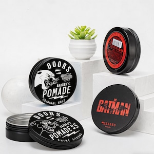 May include: Black pomade tins with white and red text and graphics. One tin features a panther graphic and the words "DOORS BARBER'S POMADE ORIGINAL HOLD". Another tin has "THE BATMAN" in red text. A small succulent plant is in a white pot.