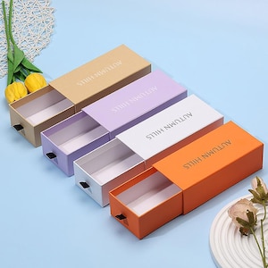 Może przedstawiać: Kolekcja prostokątnych pudełek przesuwnych w różnych kolorach: beżowym, lawendowym, białym i pomarańczowym. Każde pudełko ma białe wnętrze i czarny uchwyt. Na górze każdego pudełka wydrukowano tekst "AUTUMN HILLS".