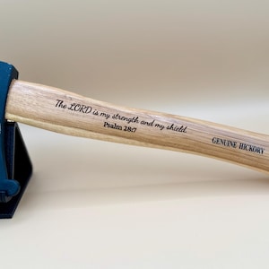 May include: A 1 1/4 lb hatchet with a matte black head and a light brown hickory handle. The head features the text Waymaker and Isaiah 42:16. The handle is engraved with The LORD is my strength and my shield. Psalm 28:7 and GENUINE HICKORY.