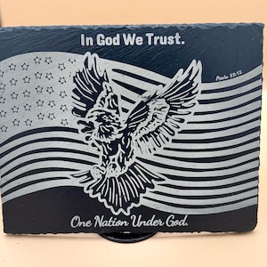 May include: Black rectangular slate coaster with an engraved American flag design, a soaring eagle, and the text "In God We Trust" and "One Nation Under God." The design is in white against the dark slate.