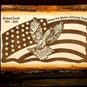 May include: A wooden plaque with an engraved American flag design and an eagle. The text reads "Now He Soars Among Eagles" and includes the name "Richard Smith 1984-2024". A candle and a potted plant are in the background.
