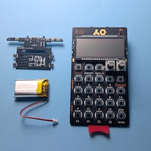 May include: A black electronic music device with a screen, buttons, and knobs, along with a circuit board and battery. The device has the text "K.O!" and "OY" printed on it. A red tab is attached to the bottom.