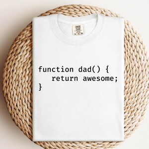 K&ouml;nnte beinhalten: Wei&szlig;es T-Shirt mit dem Text "function dad() { return awesome; }" in Schwarz, ein humorvolles Design im Coding-Stil. Das Shirt ist von der Marke Comfort Colors und wird auf einer hellbraunen, gewebten Oberfl&auml;che pr&auml;sentiert.