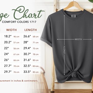 K&ouml;nnte beinhalten: Ein dunkelgraues T-Shirt h&auml;ngt an einem Holzb&uuml;gel, mit einer Gr&ouml;&szlig;entabelle links. Die Tabelle zeigt Ma&szlig;e in Zoll und Zentimetern f&uuml;r verschiedene Gr&ouml;&szlig;en, einschlie&szlig;lich Breite und L&auml;nge. Der Text "Size Chart" steht oben.