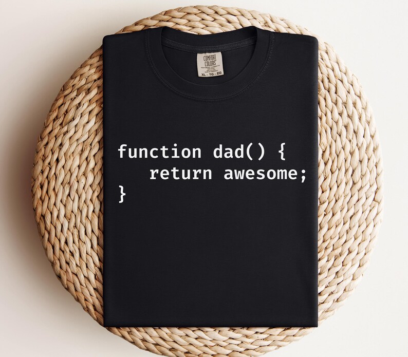 K&ouml;nnte beinhalten: Schwarzes T-Shirt mit dem wei&szlig;en Text "function dad() { return awesome; }". Das gefaltete T-Shirt liegt auf einer runden, gewebten Matte. Das Shirt hat einen Rundhalsausschnitt und kurze &Auml;rmel.