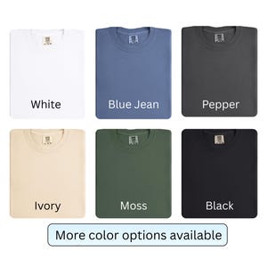K&ouml;nnte beinhalten: Gefaltete T-Shirts in den Farben Wei&szlig;, Blue Jean, Pfeffer, Elfenbein, Moos und Schwarz. Jedes Shirt hat ein Etikett am Halsausschnitt. Der Text "More color options available" ist unten im Bild zu sehen.