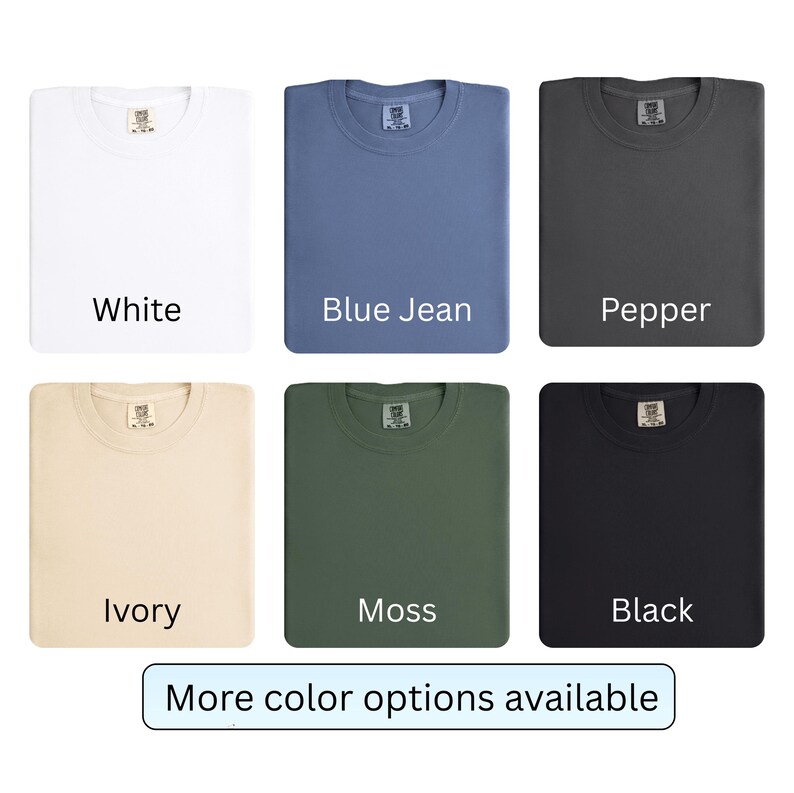 K&ouml;nnte beinhalten: Gefaltete T-Shirts in den Farben Wei&szlig;, Blue Jean, Pfeffer, Elfenbein, Moos und Schwarz. Jedes Shirt hat ein Etikett am Halsausschnitt. Der Text "More color options available" ist unten im Bild zu sehen.