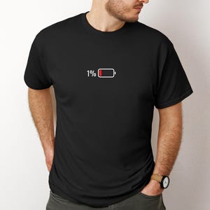 May include: Black crew neck t-shirt with a minimalist graphic of a battery at 1% charge. The battery icon is red and white, and the number "1%" is printed above it. The shirt is a casual, everyday wear item.