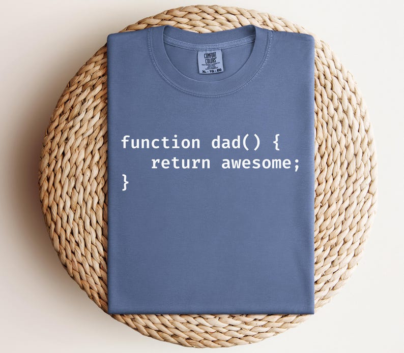K&ouml;nnte beinhalten: Ein blaues T-Shirt mit dem Text "function dad() { return awesome; }" in Wei&szlig;. Das T-Shirt ist gefaltet und auf einer gewebten, kreisf&ouml;rmigen Oberfl&auml;che platziert. Das T-Shirt hat einen Rundhalsausschnitt und kurze &Auml;rmel.