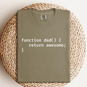 K&ouml;nnte beinhalten: Olivgr&uuml;nes T-Shirt mit dem wei&szlig;en Text "function dad() { return awesome; }". Das T-Shirt wird auf einer gewebten, kreisf&ouml;rmigen Oberfl&auml;che pr&auml;sentiert.